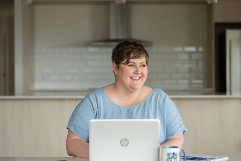 A photograph of Melanie smiling. She has dark blonde hair and is wearing a blue shirt. She is sitting behind a laptop at a desk.